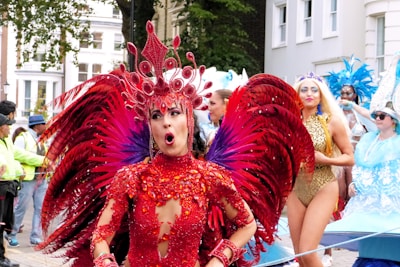 A vibrant street parade featuring performers in elaborate costumes. A person in a stunning red costume with large feathered wings and an intricate headpiece stands prominently. Surrounding them are other performers in equally dazzling outfits of gold and blue with feathers and sequins. The background shows a street lined with buildings and onlookers.