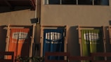 Three fabric banners hang against a beige wall, each featuring the logo of 'East West Brewing Company' and the text 'Made in Vietnam'. The banners are in three different colors: orange, blue, and green. The sunlight casts shadows, enhancing the textures and colors.