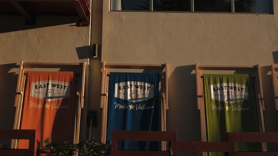 Three fabric banners hang against a beige wall, each featuring the logo of 'East West Brewing Company' and the text 'Made in Vietnam'. The banners are in three different colors: orange, blue, and green. The sunlight casts shadows, enhancing the textures and colors.