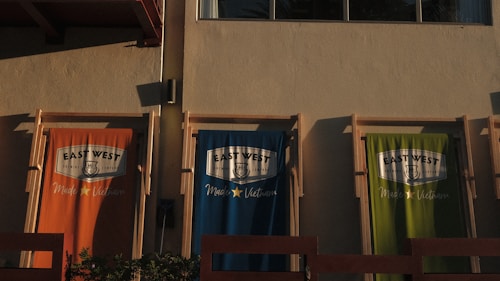 Three fabric banners hang against a beige wall, each featuring the logo of 'East West Brewing Company' and the text 'Made in Vietnam'. The banners are in three different colors: orange, blue, and green. The sunlight casts shadows, enhancing the textures and colors.