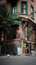 A corner building with an upper floor featuring green painted window frames and a small balcony. Below, a small doner kebab shop with illuminated signage displays images of food. Two people are standing near the entrance of the shop, and a tree is next to the sidewalk.