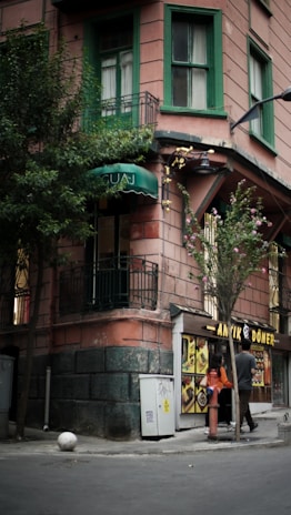 A corner building with an upper floor featuring green painted window frames and a small balcony. Below, a small doner kebab shop with illuminated signage displays images of food. Two people are standing near the entrance of the shop, and a tree is next to the sidewalk.