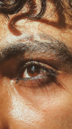 A close-up of a person's eye with thick eyebrows and natural skin texture. The lighting highlights the glistening skin and rich brown color of the eye, with a soft curl of hair above the brow.
