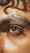 A close-up of a person's eye with thick eyebrows and natural skin texture. The lighting highlights the glistening skin and rich brown color of the eye, with a soft curl of hair above the brow.