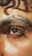 A close-up of a person's eye with thick eyebrows and natural skin texture. The lighting highlights the glistening skin and rich brown color of the eye, with a soft curl of hair above the brow.