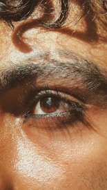 A close-up of a person's eye with thick eyebrows and natural skin texture. The lighting highlights the glistening skin and rich brown color of the eye, with a soft curl of hair above the brow.