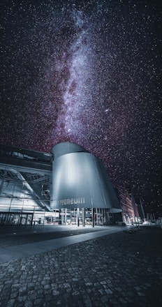A modern architectural building with a cylindrical metallic structure stands against a breathtaking night sky filled with countless stars and a clearly visible Milky Way. The ground is paved with cobblestones, adding texture and depth to the foreground.