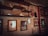 A rustic room features vintage-style shelves with retro decor items such as old radios, figurines, and bottles. Framed posters with vintage graphics and writing adorn the worn walls, adding a nostalgic touch. The atmosphere is dimly lit, emphasizing the aged and cozy tone.