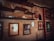 A rustic room features vintage-style shelves with retro decor items such as old radios, figurines, and bottles. Framed posters with vintage graphics and writing adorn the worn walls, adding a nostalgic touch. The atmosphere is dimly lit, emphasizing the aged and cozy tone.