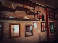 A rustic room features vintage-style shelves with retro decor items such as old radios, figurines, and bottles. Framed posters with vintage graphics and writing adorn the worn walls, adding a nostalgic touch. The atmosphere is dimly lit, emphasizing the aged and cozy tone.