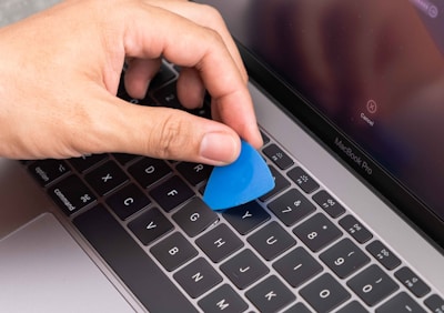 A hand is holding a blue plastic tool, possibly a pry tool or guitar pick, over the keyboard of a silver MacBook Pro laptop. The screen of the laptop is turned on, displaying a cancel button.