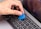 A hand is holding a blue plastic tool, possibly a pry tool or guitar pick, over the keyboard of a silver MacBook Pro laptop. The screen of the laptop is turned on, displaying a cancel button.