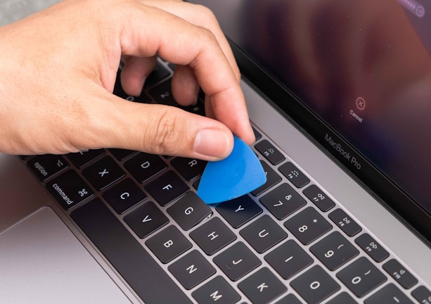 A hand is holding a blue plastic tool, possibly a pry tool or guitar pick, over the keyboard of a silver MacBook Pro laptop. The screen of the laptop is turned on, displaying a cancel button.