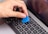 A hand is holding a blue plastic tool, possibly a pry tool or guitar pick, over the keyboard of a silver MacBook Pro laptop. The screen of the laptop is turned on, displaying a cancel button.