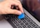 A hand is holding a blue plastic tool, possibly a pry tool or guitar pick, over the keyboard of a silver MacBook Pro laptop. The screen of the laptop is turned on, displaying a cancel button.