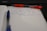 A notebook with lined paper is open, showing handwritten notes in blue ink. A blue pen is lying across the pages, and a red pen with a visible brand name is partially seen above it. The notes mention providing project schedules and deadlines and preparing a PPT for a public meeting scheduled for September 14th.