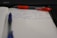 A notebook with lined paper is open, showing handwritten notes in blue ink. A blue pen is lying across the pages, and a red pen with a visible brand name is partially seen above it. The notes mention providing project schedules and deadlines and preparing a PPT for a public meeting scheduled for September 14th.