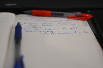 A notebook with lined paper is open, showing handwritten notes in blue ink. A blue pen is lying across the pages, and a red pen with a visible brand name is partially seen above it. The notes mention providing project schedules and deadlines and preparing a PPT for a public meeting scheduled for September 14th.