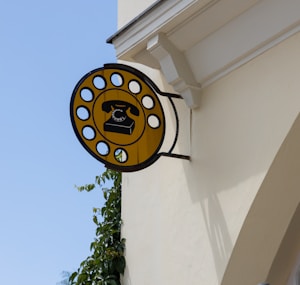 A vintage-style sign depicting a rotary dial telephone, mounted on the side of a building. The sign is circular with a bright yellow background and black outlines, designed to resemble a rotary dial. It is fixed to a light-colored wall, with some green foliage visible climbing up the structures. The sky is clear with a pale blue hue.