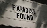 Black bold letters spell out 'Paradise Found' on a vintage, lightbox-style sign. The background is a soft, blurred gradient, giving a nostalgic and slightly worn appearance.
