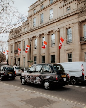 A large, neoclassical building with multiple Canadian flags hanging from its facade. The building appears to be an embassy or government office, and is situated along a street lined with several black cabs and a white van. The architecture is grand and symmetrical, with tall columns supporting the entrance. Leafless trees and cloudy skies suggest it may be winter or early spring.