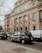 A large, neoclassical building with multiple Canadian flags hanging from its facade. The building appears to be an embassy or government office, and is situated along a street lined with several black cabs and a white van. The architecture is grand and symmetrical, with tall columns supporting the entrance. Leafless trees and cloudy skies suggest it may be winter or early spring.