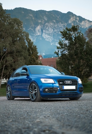 A blue Audi car is parked on gravel with a backdrop of lush green trees and majestic mountains. The car has sleek lines and modern design elements, including distinctive alloy wheels and a prominent front grille.