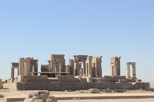 Ancient ruins of a once-grand stone structure featuring large columns and arches, set against a clear blue sky. The site appears weathered by time but retains a majestic and historic aura.