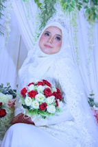 A bride wearing a white hijab and intricate lace gown holds a vibrant bouquet of red and white roses. She is adorned with a sparkling tiara and surrounded by lush greenery and delicate drapery.