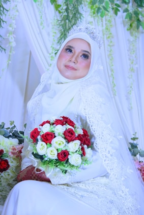 A bride wearing a white hijab and intricate lace gown holds a vibrant bouquet of red and white roses. She is adorned with a sparkling tiara and surrounded by lush greenery and delicate drapery.