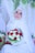A bride wearing a white hijab and intricate lace gown holds a vibrant bouquet of red and white roses. She is adorned with a sparkling tiara and surrounded by lush greenery and delicate drapery.