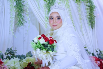 A bride dressed in a white hijab and gown adorned with lace details is holding a bouquet of red and white roses. She is wearing a silver tiara and is surrounded by floral decorations with draped white fabric and green leafy vines.
