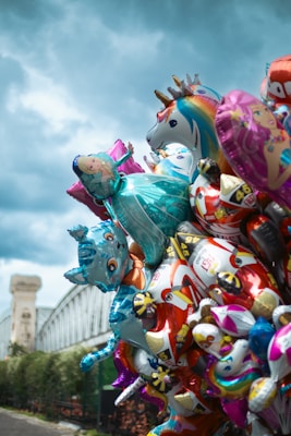 A cluster of colorful helium balloons, featuring various cartoon characters and shapes, including a unicorn, several animals, and a princess. The balloons are gathered together against an outdoor background, which includes part of a bridge and an overcast sky.