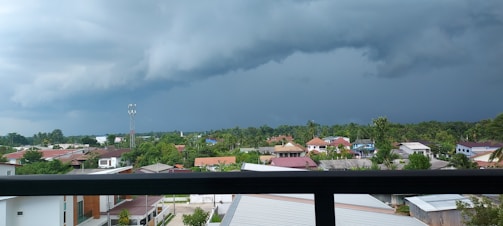A panoramic view of a suburban area with a line of houses against the backdrop of a dark and stormy sky. Dense green vegetation surrounds the community, and a tall communication tower is visible. Large, ominous clouds loom overhead, suggesting an impending storm.