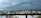 A panoramic view of a suburban area with a line of houses against the backdrop of a dark and stormy sky. Dense green vegetation surrounds the community, and a tall communication tower is visible. Large, ominous clouds loom overhead, suggesting an impending storm.