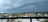 A panoramic view of a suburban area with a line of houses against the backdrop of a dark and stormy sky. Dense green vegetation surrounds the community, and a tall communication tower is visible. Large, ominous clouds loom overhead, suggesting an impending storm.