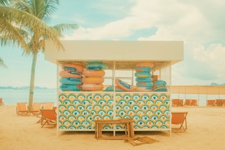 A beach scene featuring a colorful kiosk filled with neatly stacked inflatable rings. The structure is decorated with vibrant patterns, and a palm tree provides a tropical feel. Sun loungers are arranged on the sandy beach, facing the calm waters under a partly cloudy sky.
