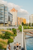 A riverside walkway lined with green and golden grasses curves along a modern urban area. People are strolling along the path, some with strollers, while a wide river runs parallel. The background features tall residential buildings with varied architectural styles, and fluffy clouds drift across the blue sky.