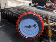 A close-up view of a pressure gauge with a red casing, displaying measurements in psi and kPa. The gauge is connected to brass fittings and placed against a mechanical background with various tubes and metallic components.