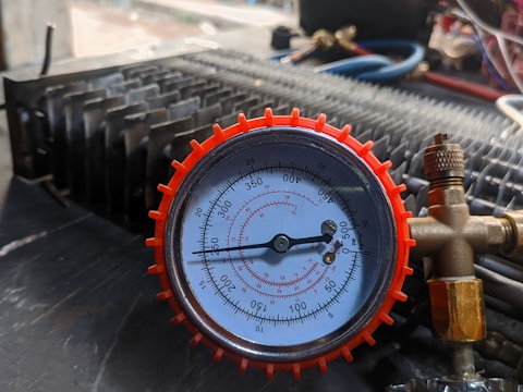 A close-up view of a pressure gauge with a red casing, displaying measurements in psi and kPa. The gauge is connected to brass fittings and placed against a mechanical background with various tubes and metallic components.