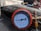 A close-up view of a pressure gauge with a red casing, displaying measurements in psi and kPa. The gauge is connected to brass fittings and placed against a mechanical background with various tubes and metallic components.