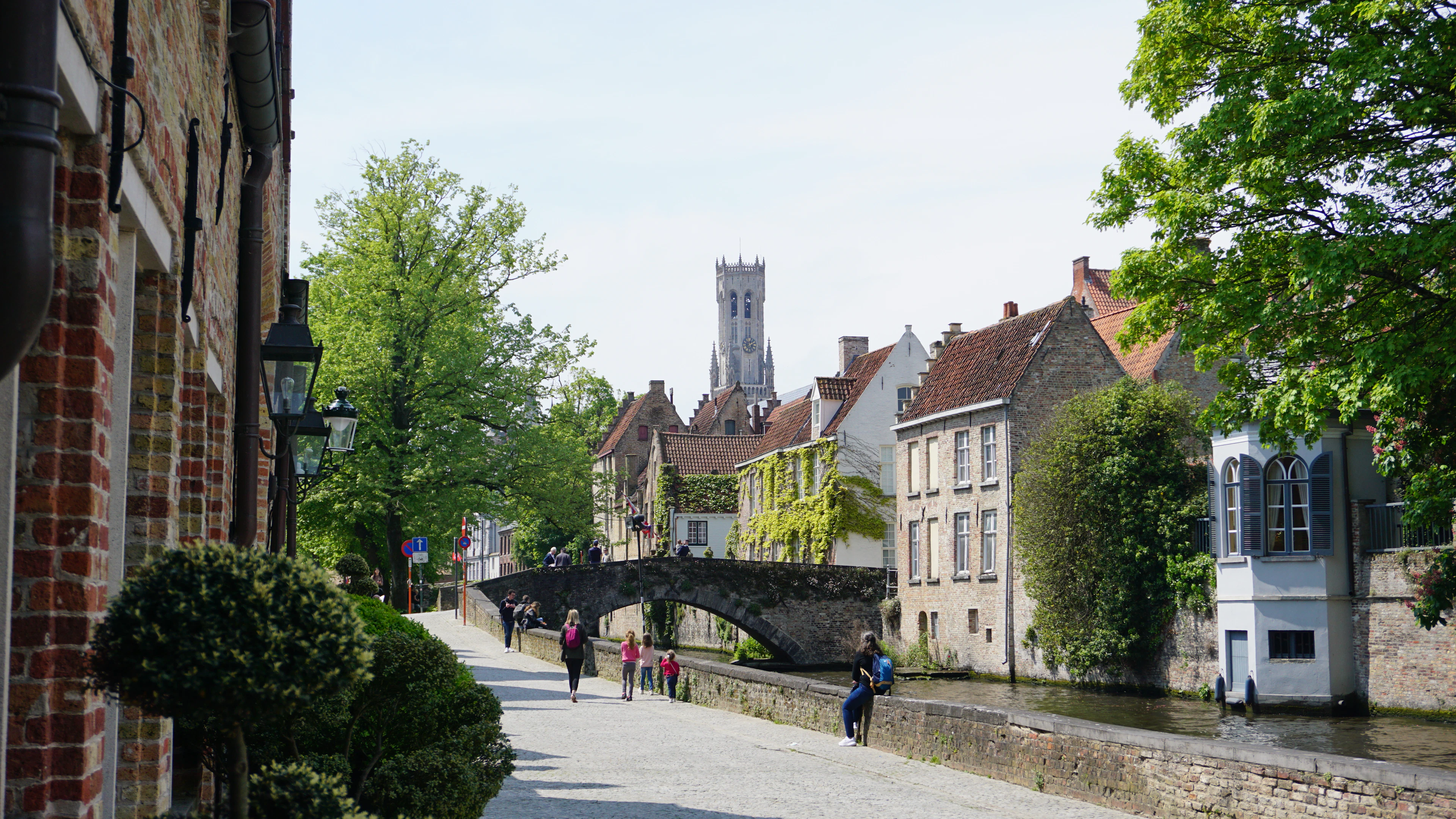 A stone bridge over a Bruges canal with a church spire rising above spring greenery in the background.