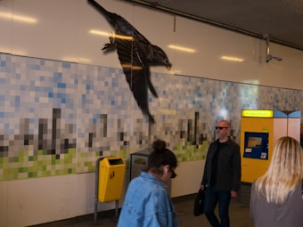 A mosaic-style wall art features large pixelated shapes resembling a cityscape. A realistic black bird is depicted in flight across the wall. Below, people are walking through the space, including a man wearing sunglasses and holding a bag, and a woman with sunglasses on her head. A yellow mailbox and a digital machine or screen are visible to the right.