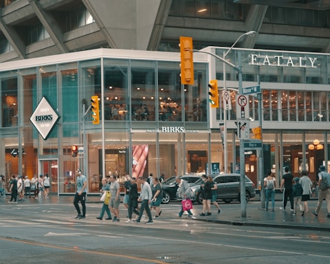 A busy urban street scene with a modern glass-fronted building housing retail stores like Birks and Eataly. Pedestrians are crossing the street, and the area is bustling with activity. Traffic lights and street signs are visible against the backdrop of the tall concrete structure.