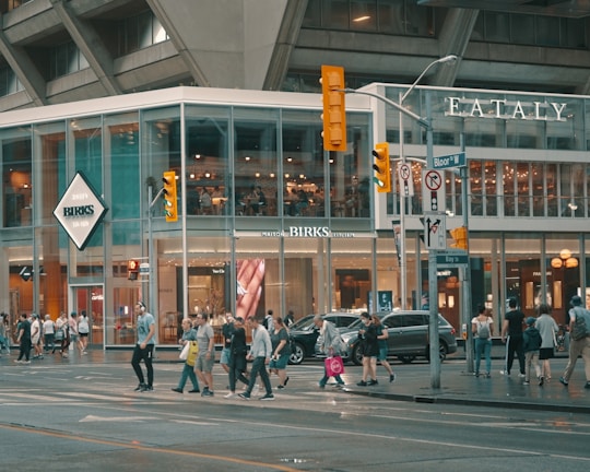 A busy urban street scene with a modern glass-fronted building housing retail stores like Birks and Eataly. Pedestrians are crossing the street, and the area is bustling with activity. Traffic lights and street signs are visible against the backdrop of the tall concrete structure.