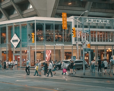 A busy urban street scene with a modern glass-fronted building housing retail stores like Birks and Eataly. Pedestrians are crossing the street, and the area is bustling with activity. Traffic lights and street signs are visible against the backdrop of the tall concrete structure.
