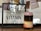 A framed sign with the text 'Coffee, The Other Vitamin C' is displayed on a wooden surface. Next to it is a layered drink in a transparent glass jar. The drink has distinct layers of black, cream, and pink, topped with a frothy purple layer. Behind, there are blurred photos with coffee-themed illustrations.