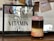 A framed sign with the text 'Coffee, The Other Vitamin C' is displayed on a wooden surface. Next to it is a layered drink in a transparent glass jar. The drink has distinct layers of black, cream, and pink, topped with a frothy purple layer. Behind, there are blurred photos with coffee-themed illustrations.