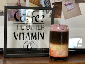 A framed sign with the text 'Coffee, The Other Vitamin C' is displayed on a wooden surface. Next to it is a layered drink in a transparent glass jar. The drink has distinct layers of black, cream, and pink, topped with a frothy purple layer. Behind, there are blurred photos with coffee-themed illustrations.