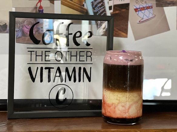 A framed sign with the text 'Coffee, The Other Vitamin C' is displayed on a wooden surface. Next to it is a layered drink in a transparent glass jar. The drink has distinct layers of black, cream, and pink, topped with a frothy purple layer. Behind, there are blurred photos with coffee-themed illustrations.